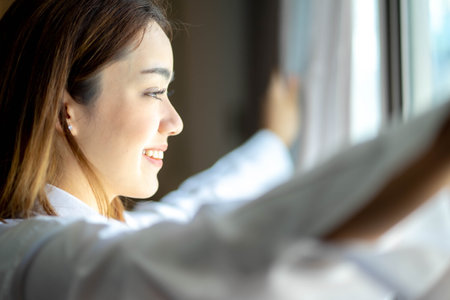 Overjoyed young beautiful woman open curtains look in window distance meet welcome new day. Smiling female feel excited about life career opportunities or perspectives.の写真素材