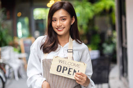Portrait of a smiling young woman in apron holding open sign board while standing at the cafe. The...