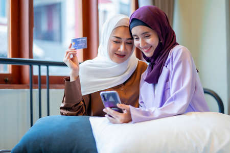 Two Muslim women wearing a hijab using a smartphone, shopping online with a credit card. muslim...
