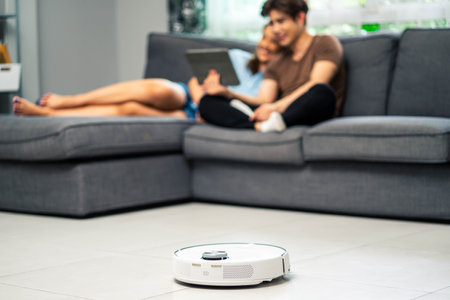 Young couple resting on the couch while robotic vacuum cleaner doing its work at home. Household robots concept.の写真素材