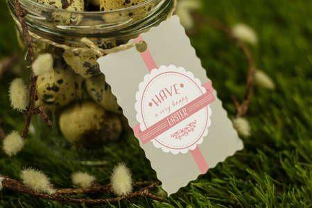 A jar full of quail eggs standing on grass with pussy willow around it and a label saying have a...