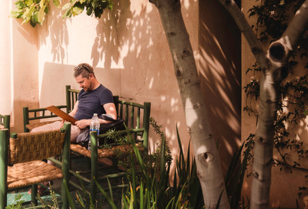 One Mid-adult, casual attire, caucasian man sat outdoors in the shade under a tree reading menu, in...