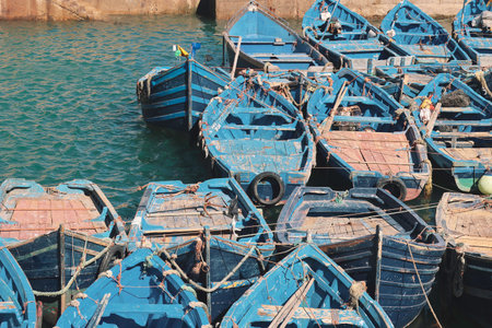 Historical port of Essaouira, Morocco with blue fishing boats docked beside famous ancient walls, capturing the coastal charm and maritime heritage of this iconic moroccan seaside destinationの写真素材