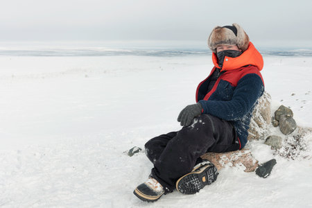 Adult caucasian male musher dressed in full arctic gear sitting on snowy tundra landscape in frozen...