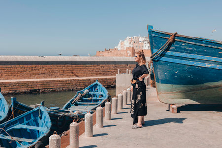 Young mid-adult female caucasian tourist standing at port in Essaouira, looking out towards the ocean, admiring the view, with traditional blue boats, ancient walled city in background, Moroccoの写真素材