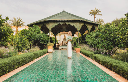 A woman in stylish casual attire, in a lush garden with green hedges and palm trees, facing a traditional Moroccan gazebo. The pathway is lined with vibrant greenery and flowers, creating a serene atmosphere.のeditorial素材