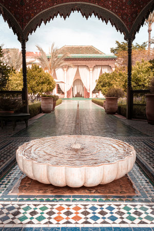 A serene courtyard featuring a large, round fountain surrounded by intricate tile work and lush greenery. The background shows elegant draped curtains and traditional Moroccan architectureのeditorial素材