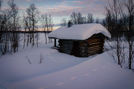 Wilderness log cabin blanketed in deep snow at dusk, twilight sky in Finnish arctic circle, remote, quiet, winter snowy Nordic landscape, Tarvantovaara Wilderness Area, Finlandのeditorial素材