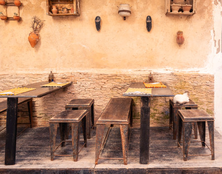 Charming rustic outdoor cafe restaurant seating with wooden tables and stools in Essaouira, Morocco, traditional decor on aged plaster wall with pottery, a white cat resting on bench, cozy atmosphereのeditorial素材