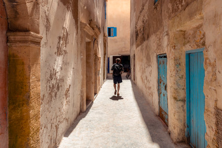 One person, young adult male walking exploring down narrow alley in Essaouira, Morocco, with aged buildings with blue painted doors, afternoon sunlight casting long shadows in historic medinaのeditorial素材