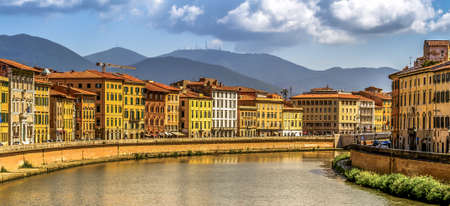 The Arno River flowing through Pisa, flanked by colorful buildings with the mountains in the backgroundの写真素材