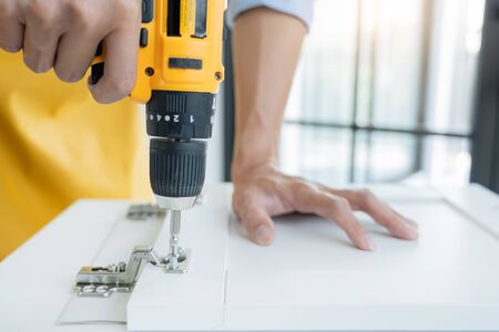 Close up portrait and details of caucasian male worker using electric screwdriver instrument in hand and repairing new wooden desk, home improvement conceptの写真素材