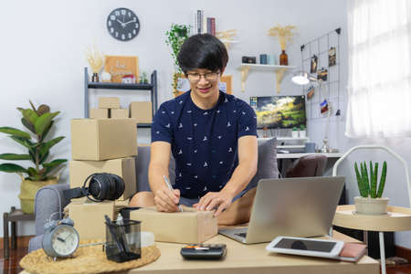 Asian man working sell online writing address on package of orders with cardboard box to send out to customer, e-commerce and online shopping conceptの写真素材