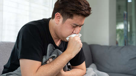 Asian man with seasonal infections Cold Blowing His Nose and sneezing into Tissue with headache lying on sofa with high fever and a flu.の写真素材