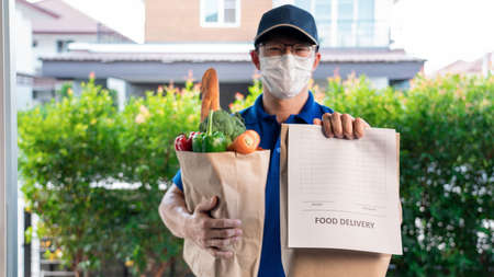 Asian deliver man in uniform with respirator face mask is delivering the paper bag waiting for the customer in front of the home during lockdown, Food delivery serviceの写真素材