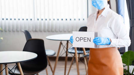 Happy waitress with protective face mask holding open sign for the reopening at her restaurant after lockdown the quarantine coronavirus or covid-19 adapt to new normalの写真素材