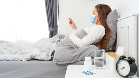 Sick Asian woman with protective mask lying in bed with high fever and measuring body temperature by thermometer and touching her forehead at her homeの写真素材