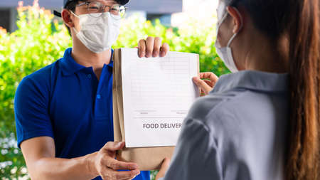 Asian deliver man wearing face mask in uniform handling bag of food and fruit give to woman costumer in front of the house, Express delivery service quarantine and shopping concept.の写真素材