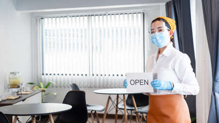 Happy waitress with protective face mask holding open sign for the reopening at her restaurant after lockdown the quarantine coronavirus or covid-19 adapt to new normalの写真素材