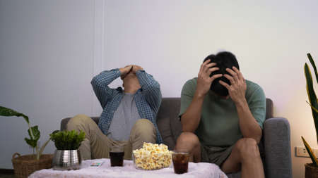 Asian friends or football fans sitting on sofa watching soccer or football game on tv at home, sports and entertainment concept.の写真素材
