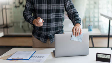Covid-19 concept a businessman cleaning his laptop to prevent Coronavirus outbreak (COVID-19) by using alcohol spray at his workplace.の写真素材