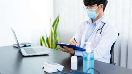 Medical concept a male doctor wearing a mask writing prescription to patient at his modern clinic...