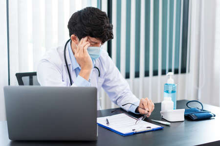 Medical concept a male doctor wearing a mask having a headache while his hard working caused by for virus outbreak  at his modern clinic.の写真素材