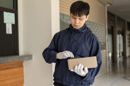 Parcel delivery concept the young mailman in navy blue coat using his smart phone to look for his customerâs address to send the parcel.の写真素材