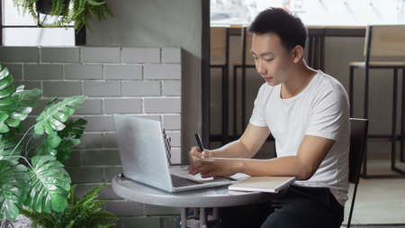 Online studying concept the young man in simple white T-shirt and black trousers working by using his laptop to search information according to the brief concepts written down on the notebook.の写真素材