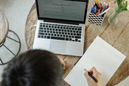 Online studying concept a black-haired student doing his homework by searching information on the internet and using this black pen to write the conclusion of his research on the paper.の写真素材