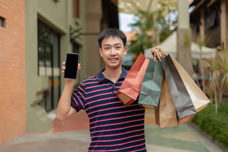 shopping concept The guy with a good mood on facial expression carrying his cell phone on his right and many shopping bags on his left.の写真素材