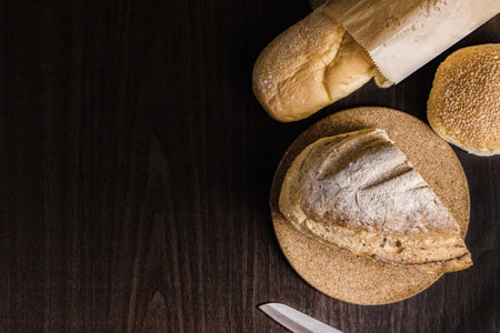 bread concept a half loaf of bread on the wooden plate, a loaf of bread into a paper bag, a knife, and a bun put on a black wooden scene.の写真素材