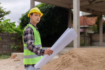 engineer concept The constructing manipulator who wears a hard yellow hat, dark-toned plaid blouse and soft green vest looking at the processing building construction.の写真素材