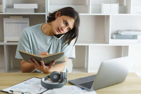 Distance education concept, Teen boy talking on phone and take note while listening lecture online.の写真素材