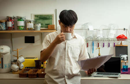 Remote work concept, Male freelancer drinking coffee and reading business document in kitchen room.の写真素材