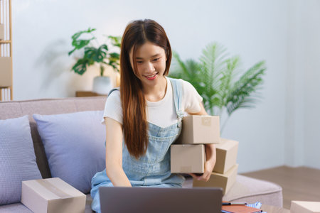 Online seller concept, Female entrepreneur holds parcel boxes and checks online orders on laptop.