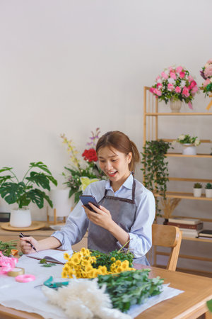 Florist concept, Woman florist looks data on smartphone to taking notes in notebook at flower shop.の写真素材