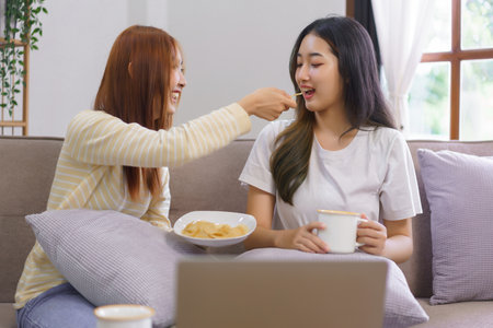 Relax at home concept, Lesbian couple drinks coffee and feeds snack to girlfriend in living room.の素材