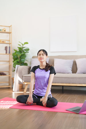 Sporty woman meditating and breathing to practice yoga exercise while learning yoga online at home.の素材