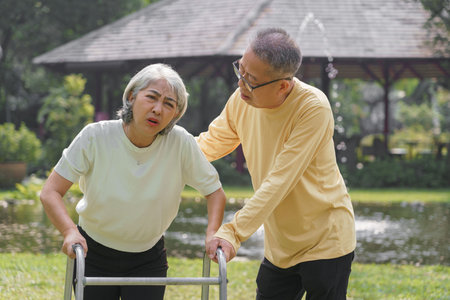 Senior man helping beloved wife walking with walker frame while doing rehab physical in the park.