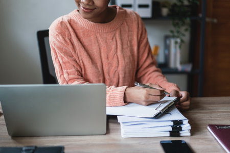 African american businesswoman reading financial data on laptop and writing notes in document.の写真素材