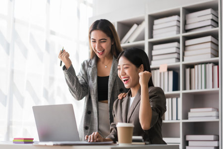 Two businesswoman reads finance data on laptop and raise arm to celebrate with successful business.