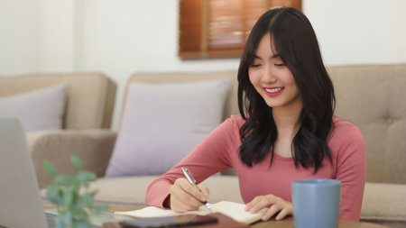 Young businesswoman reading data on laptop and writing notes in notebook while working at home.