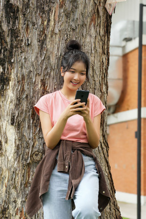 A high school student leans against a tree, smiling as she looks at her phone. The school's red brick exterior is visible in the background.の写真素材