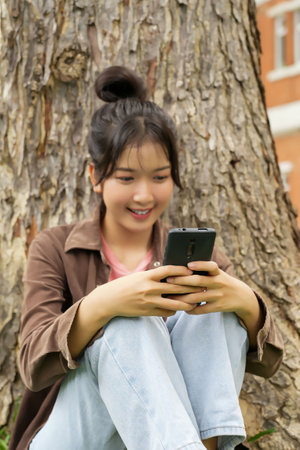A high school student is sitting on the grass under a tree, smiling as she looks at her smartphone....