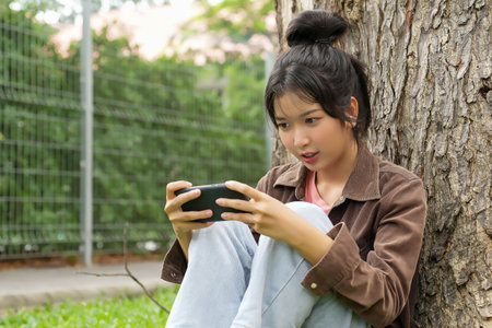 A high school student is sitting against a tree, smiling as she plays a game on her smartphone, appearing focused and entertained.の写真素材
