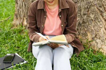 A high school student sits outside a modern school building, writing in a notebook. She is dressed casually in a brown jacket and light blue jeans, focused on her writing.の写真素材