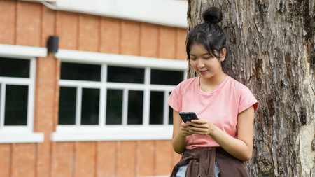 A high school student leans against a tree, smiling as she looks at her phone. The school's red brick exterior is visible in the background.の写真素材