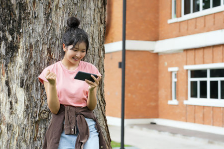 A high school student is sitting against a tree, smiling as she plays a game on her smartphone. The background shows the school's red brick exterior, enhancing the relaxedの写真素材