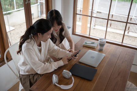 Two businesswomen using smartphone to surfing entertainment together after coworking to discussion about investment of new project and having business friendship conversation successful in workplace.の写真素材